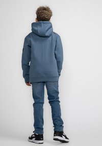 Felpa blu con un ampio cappuccio, polsini e orlo a costine. Indossata con jeans azzurri e sneakers nere. Vista da dietro.
