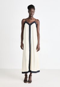 Langes, ärmelloses Kleid in Creme mit dunkelblauen Akzenten entlang des V-Ausschnitts und Saums. Hergestellt aus leichtem Stoff, mit geradem, lockeren Schnitt.