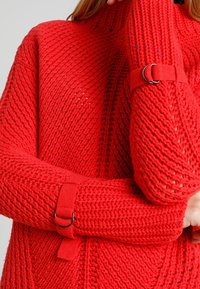 Czerwony sweter dziany z grubym splotem, prążkowanym wzorem i zapięciami na rękawach, co podkreśla jego usztywniony wygląd.