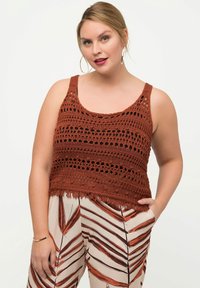 Häkeltop in Rostfarbe mit offener Maschenstruktur und Fransen unten. Kombiniert mit gemusterten Hosen mit orange-braunen Blattmotiven.