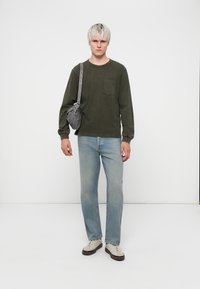 T-shirt vert à manches longues avec une poche poitrine, jeans droit bleu clair, et un sac bandoulière gris. Des baskets complètent la tenue.