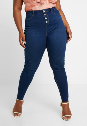 Een rond persoon draagt een donkere, blauwgekleurde skinny jeans met hoge taille en blootliggende knopen, gecombineerd met beige sandalen met hakken, en staat met gespreide benen.