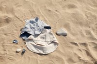 Stripet blå og hvit solhatt med øreakenter på lysfarget stoff, plassert på sandete overflate ved siden av glatte steiner.