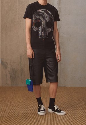 Person trägt schwarzes T-Shirt mit Design eines Schädel-Schaltkreises, schwarze Leder-Shorts, schwarze Socken, schwarze Sneakers, hält eine blau-grüne Tasche.