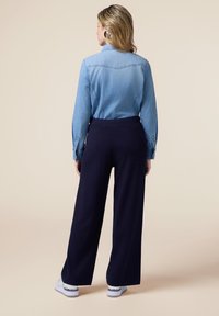 Camicia di jeans azzurra chiara con bottoni e maniche lunghe, abbinata a pantaloni larghi blu navy e sneakers bianche. Texture del tessuto lisce e visibili.