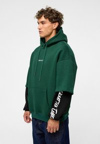 Un hoodie surdimensionné vert avec une poche kangourou, portant le mot "IMPACT" en texte blanc. Un sous-vêtement noir à manches longues arbore également un texte blanc. Design décontracté.