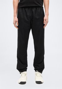 Άνδρας που φοράει χαλαρά μαύρα παντελόνια και λευκά αθλητικά παπούτσια, στέκεται με τα χέρια χαλαρά στα πλάγια σε απλό φόντο.