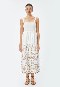 Abito bianco di cotone con corpetto elasticizzato, spalline regolabili e intricato ricamo marrone. Silhouette fluida a forma di A che arriva fino a metà polpaccio.