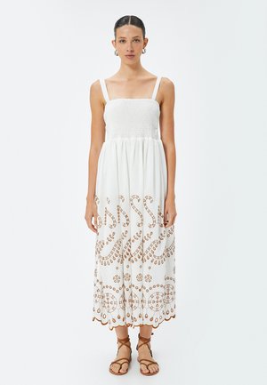 Abito bianco di cotone con corpetto elasticizzato, spalline regolabili e intricato ricamo marrone. Silhouette fluida a forma di A che arriva fino a metà polpaccio.
