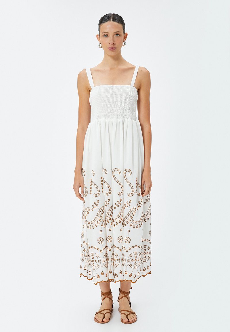 Abito bianco di cotone con corpetto elasticizzato, spalline regolabili e intricato ricamo marrone. Silhouette fluida a forma di A che arriva fino a metà polpaccio.
