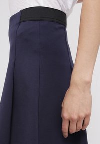 Gonna blu navy con una texture liscia, dotata di una fascia elastica nera in vita e pieghe laterali. Ingrandimento della zona vita e di una mano.