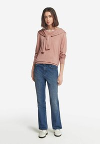 Ljusrosa tröja med en knuten detalj vid halslinjen, kombinerad med blå utsvängda jeans och vita loafers. Slätt tyg och avslappnad passform.