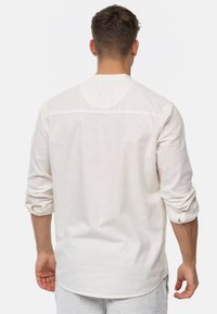 Long-sleeve shirt in light beige fabric, featuring a back yoke seam, rolled sleeves, and a relaxed fit. Textured linen material.