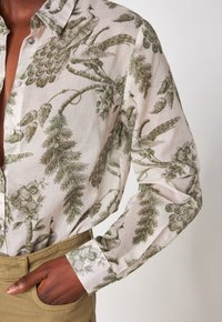 Camicia verde a fantasia con disegni di pavoni e fiori su uno sfondo chiaro, con colletto button-down e maniche lunghe.