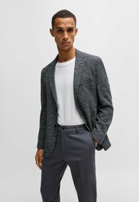 Blazer grigio scuro con texture indossato sopra una t-shirt bianca, abbinato a pantaloni grigio scuro. Vestibilità sartoriale, chiusura a due bottoni, leggero lucido.