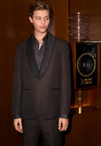 Schwarzer Anzug mit dezentem Rautenmuster, Satin-Schalkragen und passendem strukturiertem Hemd. Der Hintergrund zeigt Holzverkleidung und einen Lautsprecher.