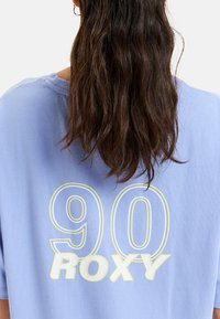 Jasnoniebieska, oversize'owa koszulka z dużą białą i żółtą grafiką "90" oraz "ROXY" na plecach. Wykonana z miękkiej bawełny, o luźnym kroju.
