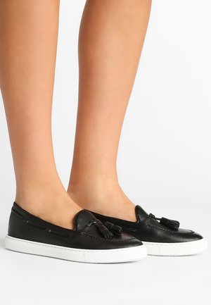 Schwarze Leder-Slipper mit Quasten, getragen an nackten Füßen, mit weißen Gummisohlen, auf weißem Hintergrund dargestellt.