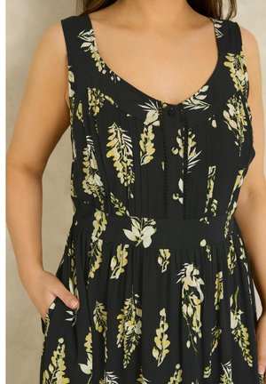 Kobieta ubrana w czarną, bezrękawną sukienkę w żółto-biały kwiatowy wzór, z ręką w bocznej kieszeni, widoczna od ramion do talii.