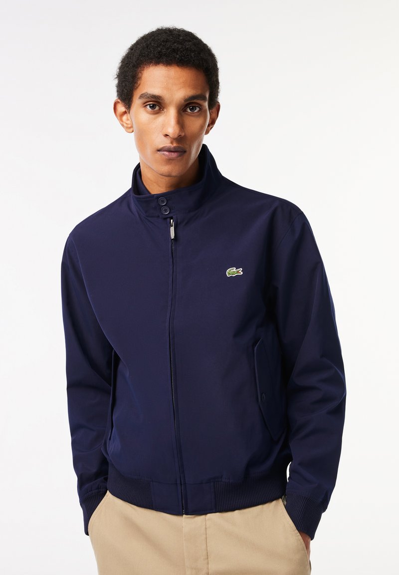 Veste bomber bleu marine avec un col montant, fermeture éclair à l'avant et poches latérales. Présente un petit logo vert sur la poitrine et des poignets côtelés.