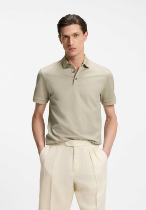 Young man with slicked-back hair wearing a light khaki textured polo shirt and cream pleated trousers, hands in pockets, neutral background.