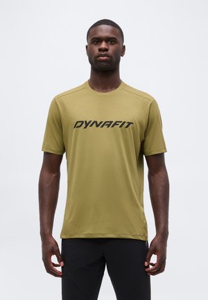 Bărbat stând cu fața înainte, purtând un tricou verde măsliniu „Dynafit” și pantaloni negri pe un fundal simplu, deschis.