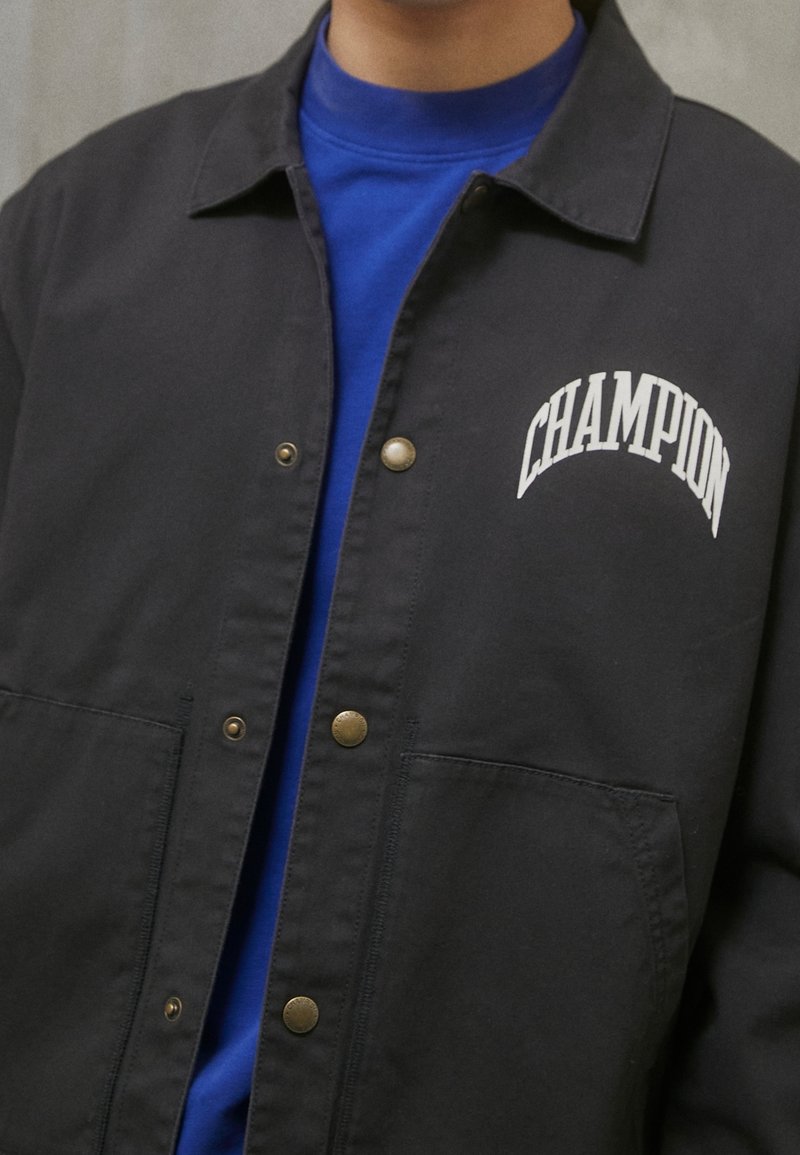 Czarna kurtka z mosiężnymi zapięciami, z haftowanym białym logo "CHAMPION" i niebieską koszulą pod spodem. Materiał nadający się do prania w pralce.