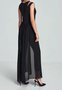 Frau in einem ärmellosen schwarzen Kleid mit durchsichtiger langer Rocküberlage und schwarzen High Heels, die seitlich vor einem einfarbigen Hintergrund steht.