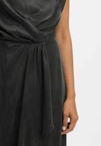 Dunkelgrünes, seidiges Kleid mit drapiertem Design. Verfügt über Falten in der Taille und eine fließende Stofftextur. Hand teilweise an der Seite sichtbar.