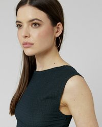 Jeune femme aux longs cheveux bruns, portant de grandes boucles d'oreilles en or et un haut noir sans manches à texture, regardant contemplativement vers la gauche.