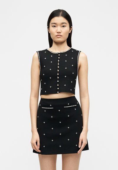 Γυναίκα που φοράει μαύρο αμάνικο κοντό τοπ και φούστα διακοσμημένα με κουμπιά από πέρλες και μικρές πέρλες σε σχήμα σταυρού.