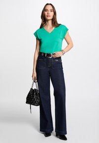 Groene v-hals t-shirt, zwarte wijde jeans met gedetailleerde zakken, riem met zilverkleurige hardware, zwarte bucket bag en zwarte puntige hakken.
