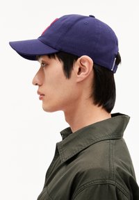 Dunkelblaue Baumwoll-Baseballkappe mit gebogenem Schirm und einem roten gestickten Logo auf der Vorderseite. Die Person trägt eine dunkelgrüne Jacke.