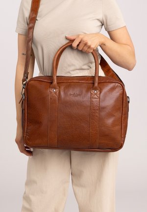 Personne tenant un sac en cuir marron de type mallette avec poignées et bandoulière, portant un pantalon beige et une chemise de couleur claire.