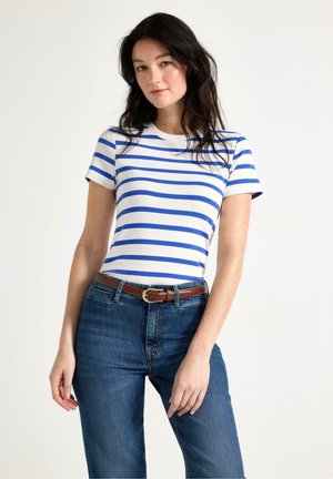 Kvinde med mørkt hår iført en hvid og blåstribet kortærmet skjorte, blå jeans og et brunt bælte mod en hvid baggrund.