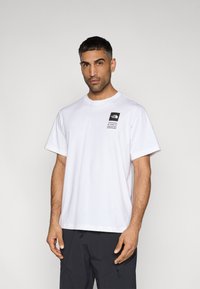 Λευκό βαμβακερό μπλουζάκι με κοντά μανίκια, στρογγυλή λαιμόκοψη και ένα μικρό μαύρο ετικέτα με λογότυπο που φέρει κείμενο στην αριστερή πλευρά του στήθους.