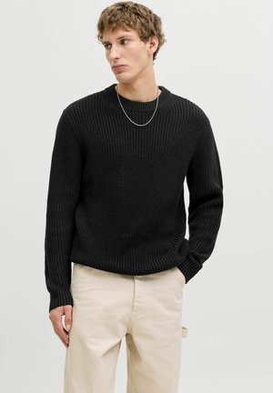 Pull en tricot noir avec un col rond et une texture côtelée, associé à un pantalon beige. Manches longues et coupe décontractée. Chaîne en argent visible.