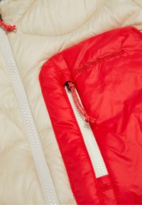 Veste isolante en matériau extérieur crème, avec une poche zippée rouge et des zips blancs avec un détail de tirage, étiquetée "Peak Performance."