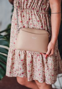 Béžová kožená crossbody taška s texturovaným povrchem, stříbrným řetízkem a vlnitým bílým logem vpředu. Pozadí se květinovým vzorem.