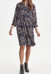 Marinblå blommig klänning med långa, genomskinliga ärmar, en V-ringning och en rynkad midja. Har en lös passform och en volangnederkant.