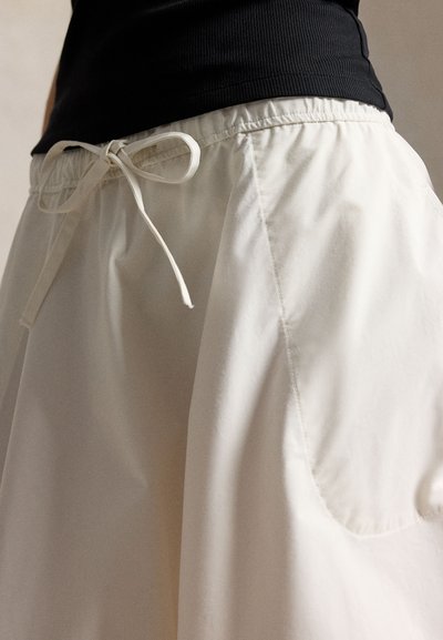 Άτομο που φοράει λευκό φαρδύ παντελόνι με κορδόνι δεμένο σε φιόγκο στη μέση και μαύρο ριμπ μπλουζάκι μέσα από το παντελόνι.