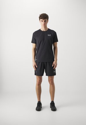 Completo sportivo nero composto da una maglietta a maniche corte e pantaloni corti, entrambi con dettagli del logo. Include sneakers nere e un design slanciato.