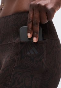 Main plaçant un smartphone noir dans la poche latérale d'un legging de sport sombre à motifs avec le logo Adidas.