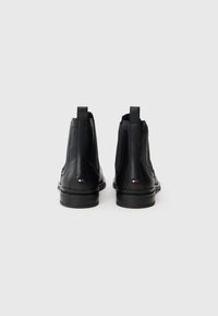Un par de botas de cuero negro hasta el tobillo con tiradores y pequeños parches en los talones en rojo, blanco y azul sobre un fondo blanco.