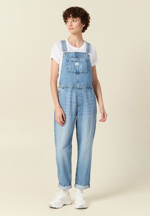 Light blue denim overalls with adjustable straps, front pocket, and rolled cuffs, paired with a white t-shirt and white sneakers.