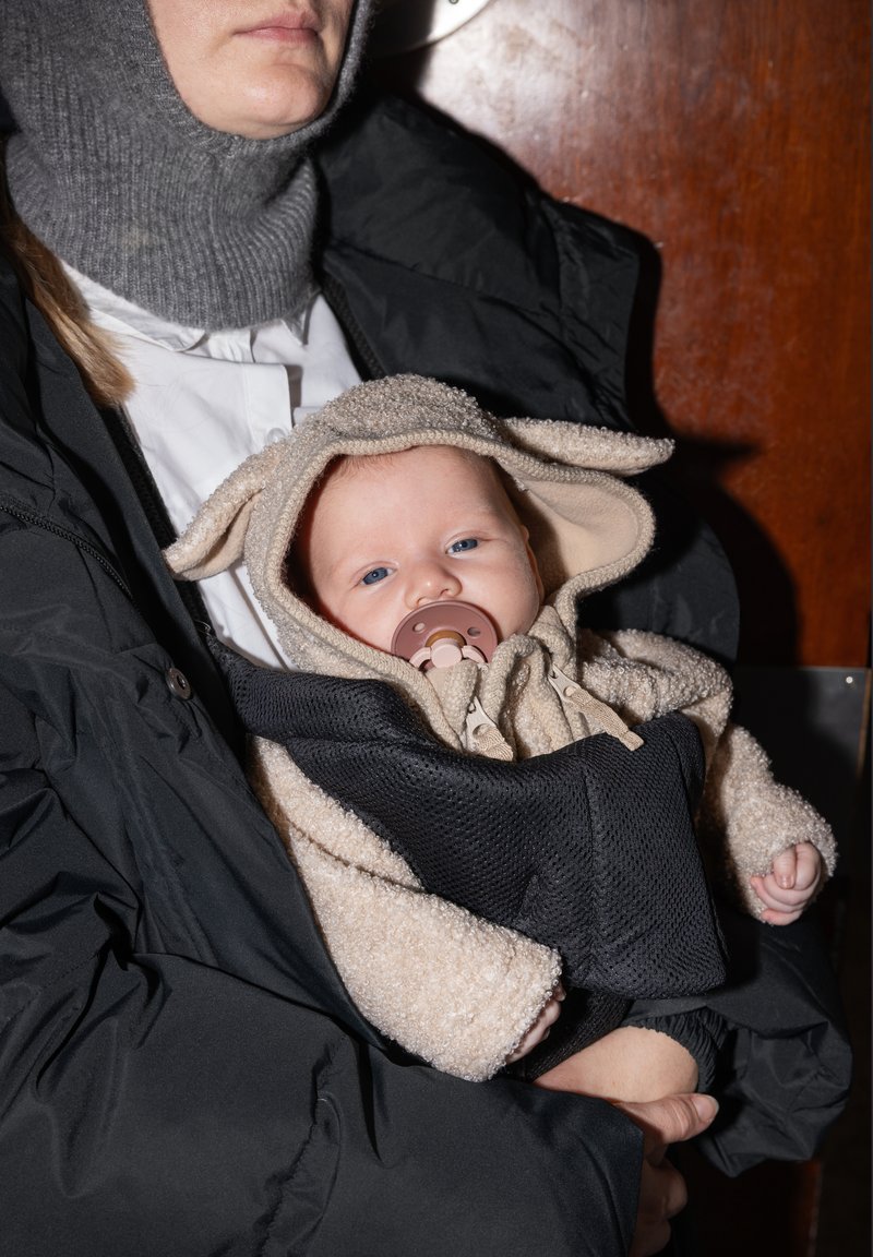 Adulte tenant un bébé vêtu d'une tenue à capuche beige avec des oreilles et une tétine, tous deux portant des vêtements d'extérieur foncés à l'intérieur.