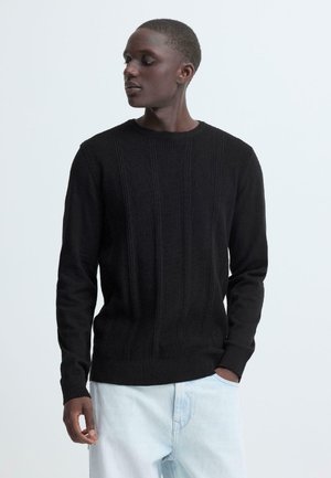Maglione nero in maglia a coste con scollo rotondo e maniche lunghe, abbinato a jeans azzurri a vestibilità rilassata, con una finitura strutturata.