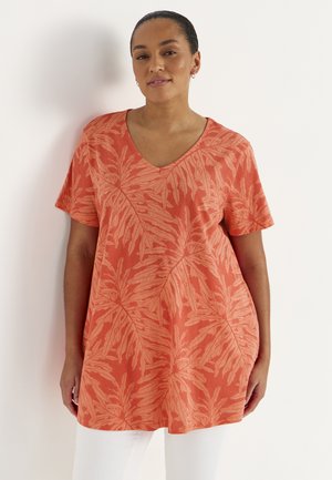 Kvinde iført en koralorange tunika med V-hals og lyse bladmønstre, stående op ad en hvid væg med afslappede hænder ned langs siderne.