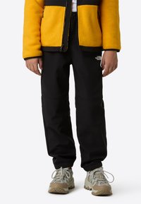 Crne softshell hlače s šavovima na koljenima, uparene sa živopisnom žutom flis jaknom. Sive i bež masivne tenisice upotpunjuju odjevnu kombinaciju.