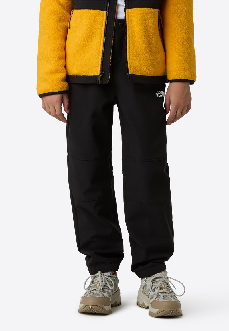 Crne softshell hlače s šavovima na koljenima, uparene sa živopisnom žutom flis jaknom. Sive i bež masivne tenisice upotpunjuju odjevnu kombinaciju.